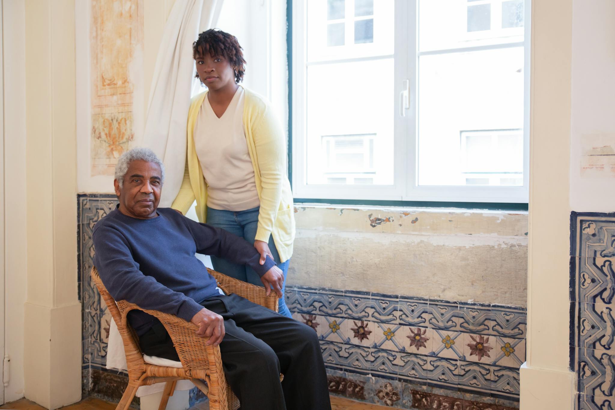 Elderly man seated with caregiver standing beside him at home in Portugal.